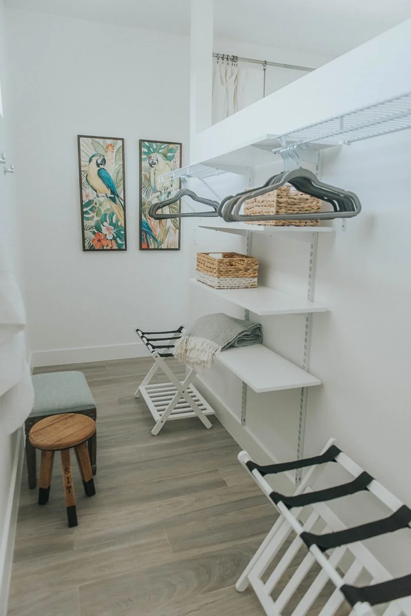 A small walk-in closet with white shelving, some wicker baskets, a folded gray blanket, a luggage rack, a small cushioned stool, and two folding chairs. There are two framed bird illustrations on the wall.