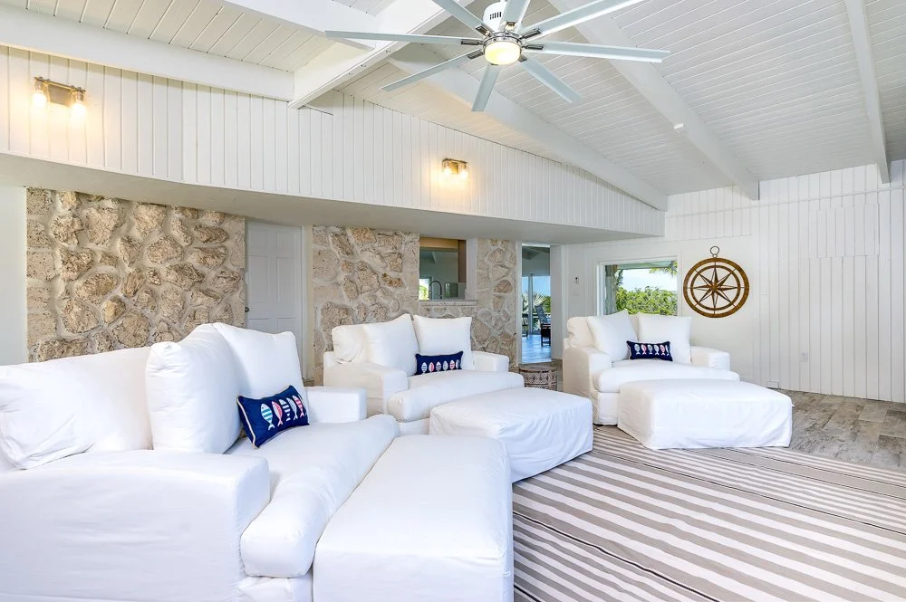Bright living room with white sofas, stone wall, white wood-paneled walls and ceiling, large window, ceiling fan, and nautical decor.