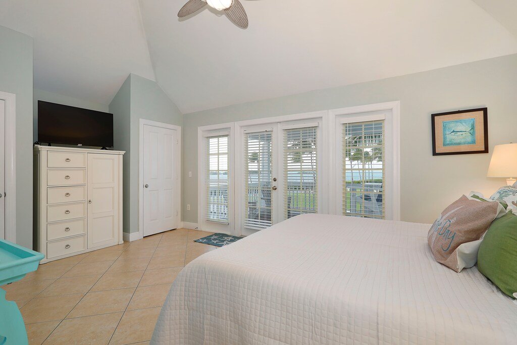 A bedroom with a white bed, a colorful pillow, a nightstand with a lamp, a piece of framed wall art, a white dresser, and a flat-screen TV on top, with glass sliding doors leading to a balcony with a view of palm trees and water.
