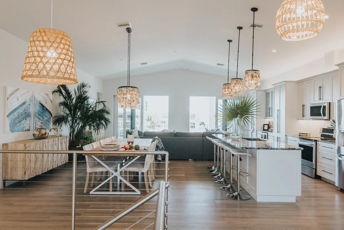 Open-concept modern kitchen and dining area with pendant lights, a large island with bar stools, a sofa in the background, and natural light from large windows.