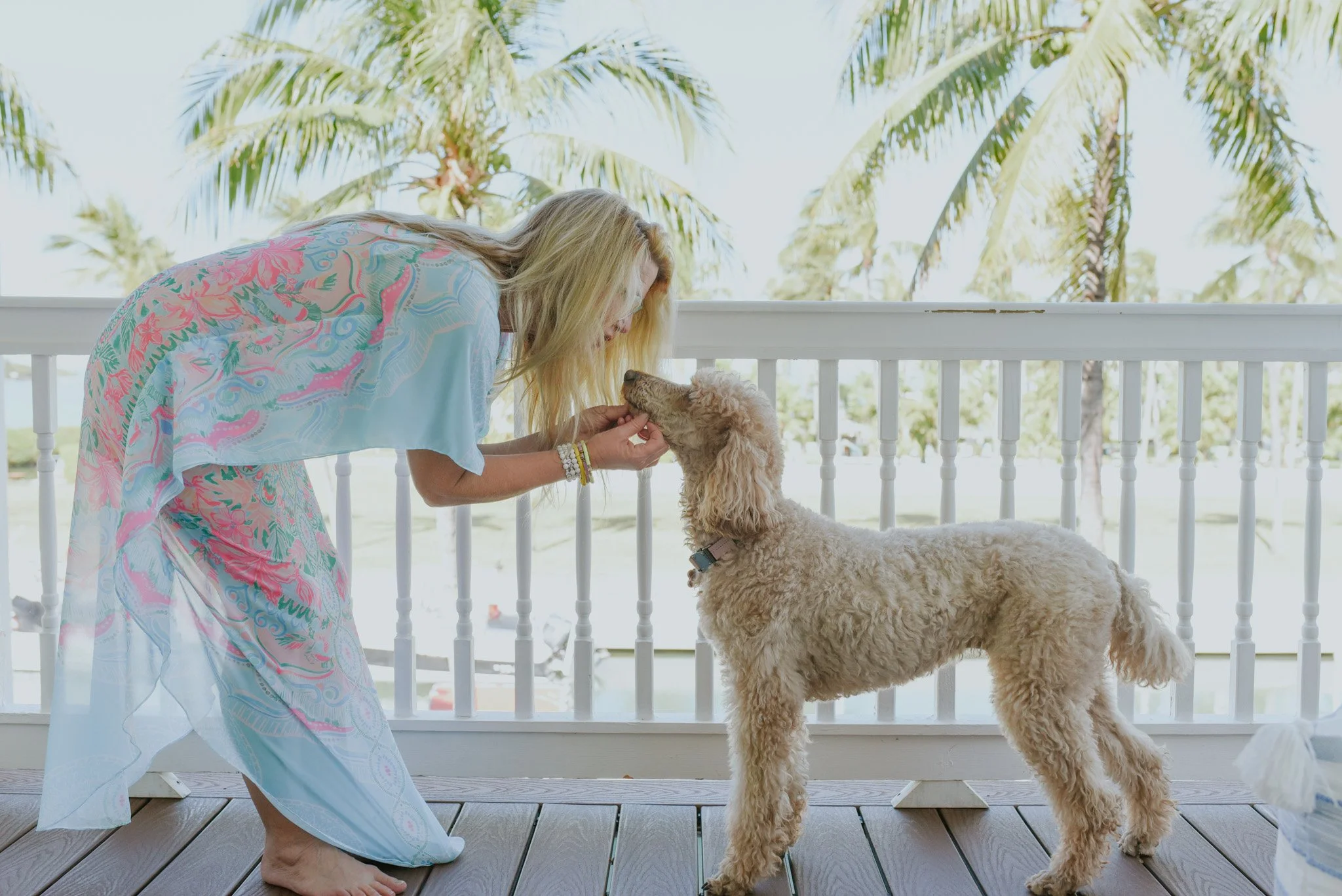 A woman with long blonde hair, wearing a pastel-colored, patterned cover-up, is bending over and gently touching a golden Cocker Spaniel puppy's nose on a wooden porch. The background features a white railing and a sunny landscape with palm trees.