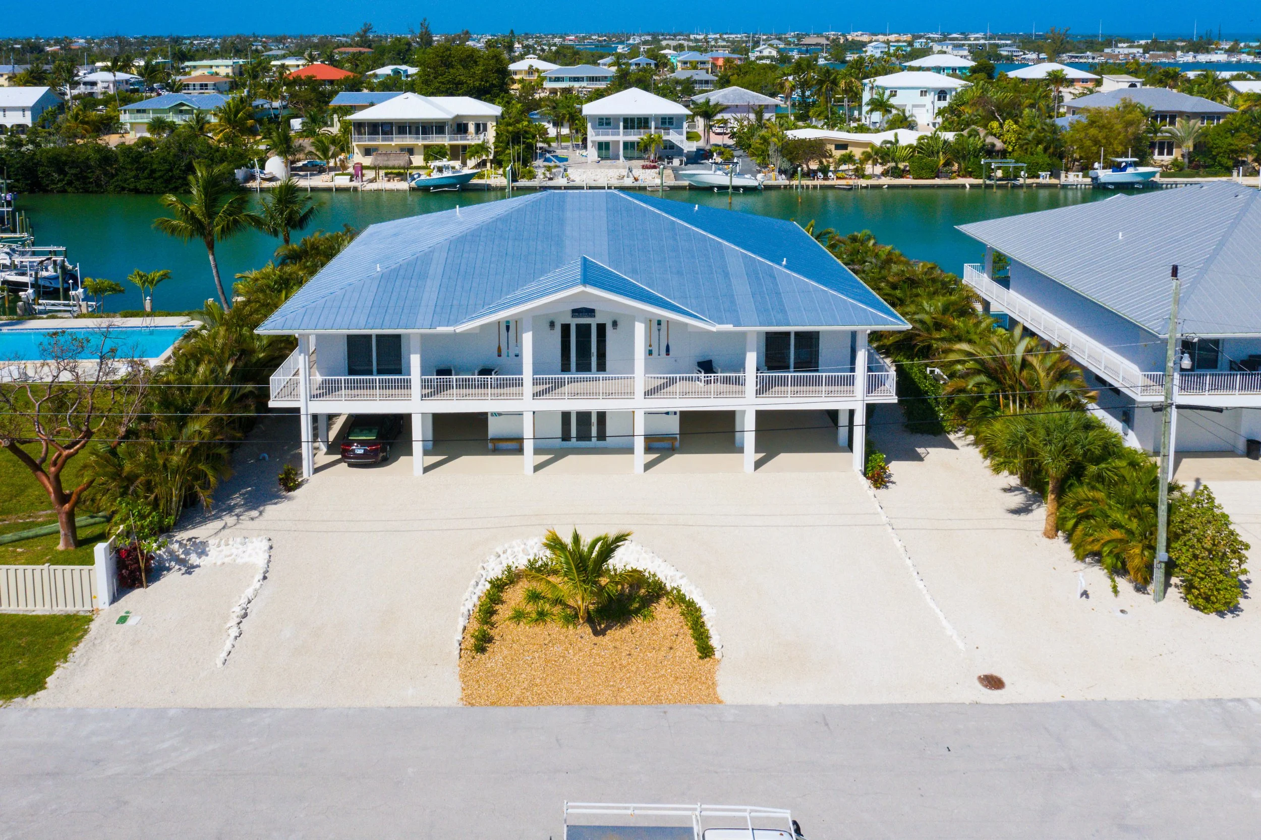 Aerial view of a modern two-story house with a light blue metal roof, white exterior, and balcony, situated on a sandy lot with a small palm tree and landscaped area in the front. The house is in a tropical neighborhood with a waterway, other similar