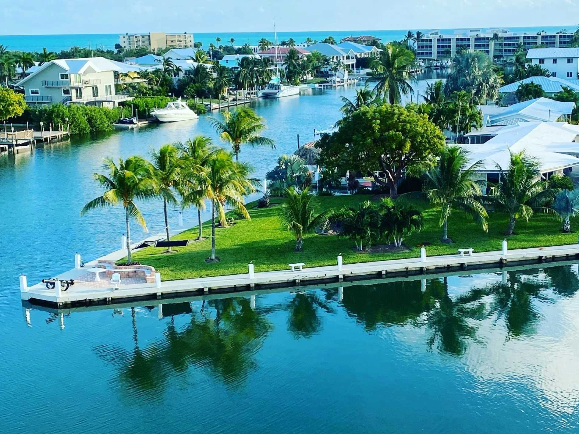 A picturesque waterfront scene with a small grassy island featuring palm trees, a white dock, and a curved stone bench. The island is surrounded by calm water, with houses and boats along the canal in the background, and the ocean in the distance und