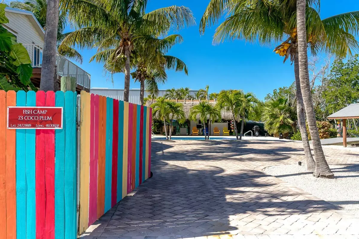 Colorful wooden fence with rainbow stripes, palm trees, a swimming pool, and a thatched-roof building in a tropical setting under a bright blue sky.