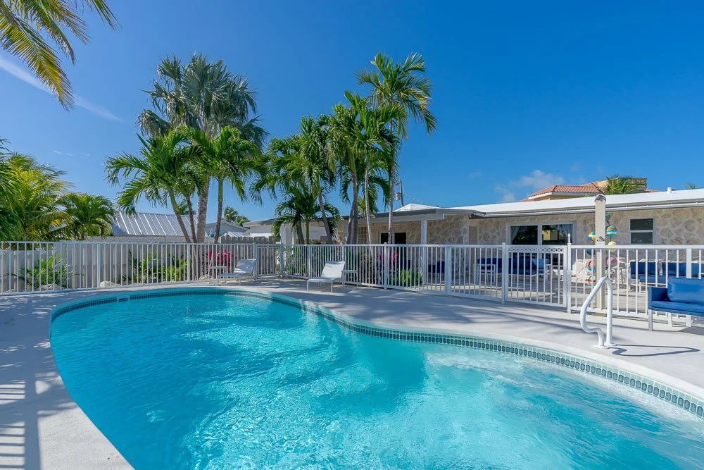 Pool with white fence, lounge chairs, palm trees, and a house under a clear blue sky.