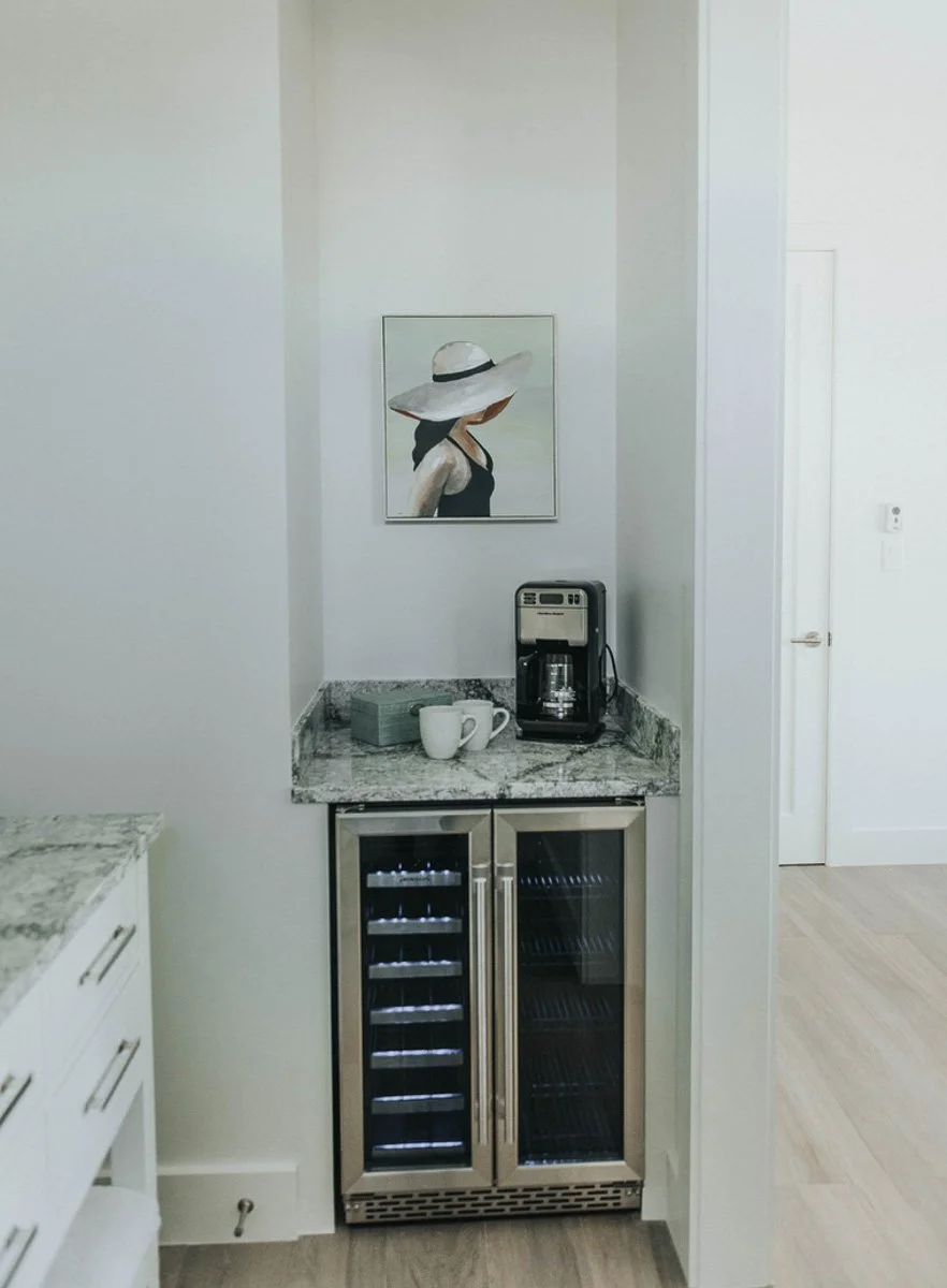A small corner of a kitchen with a coffee maker, two coffee mugs, a box, and a container on a granite countertop, with a painting of a woman wearing a wide-brimmed hat hanging on the wall above, and a wine cooler below.