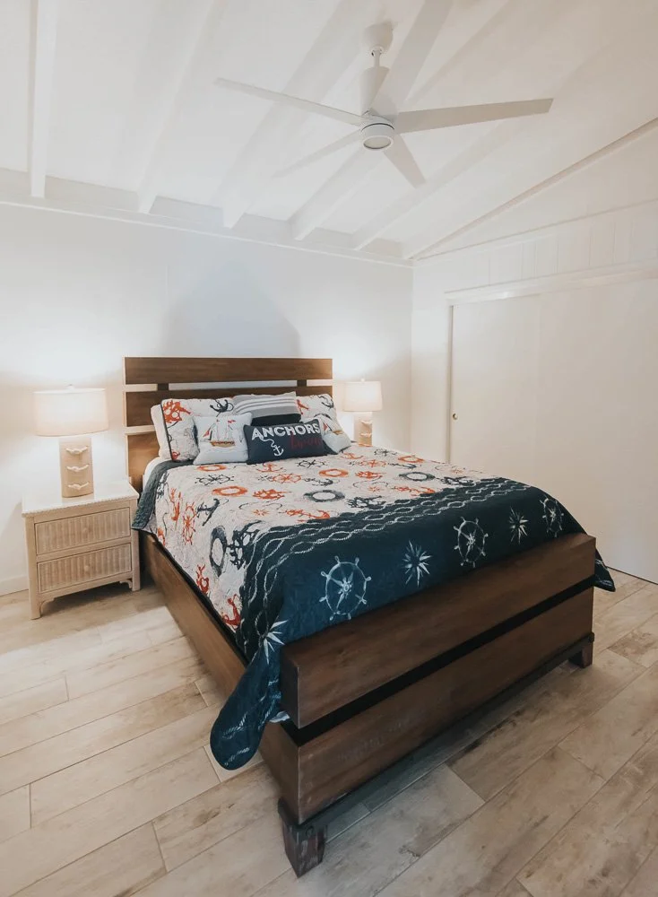 A bedroom with a wooden bed frame and a nautical-themed quilt and pillows. There are two matching nightstands with lamps on each side of the bed. The room has white walls, a white ceiling with exposed beams, and a ceiling fan. The floor is light-colo