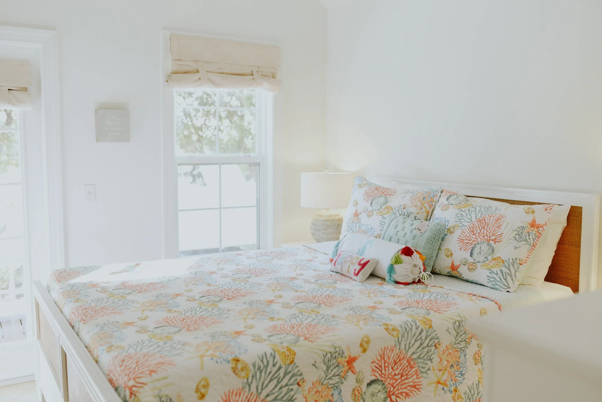 A bedroom with a white bed featuring ocean-themed bedding, pillows, and a throw, next to two windows with beige Roman shades, and a nightstand with a lamp.