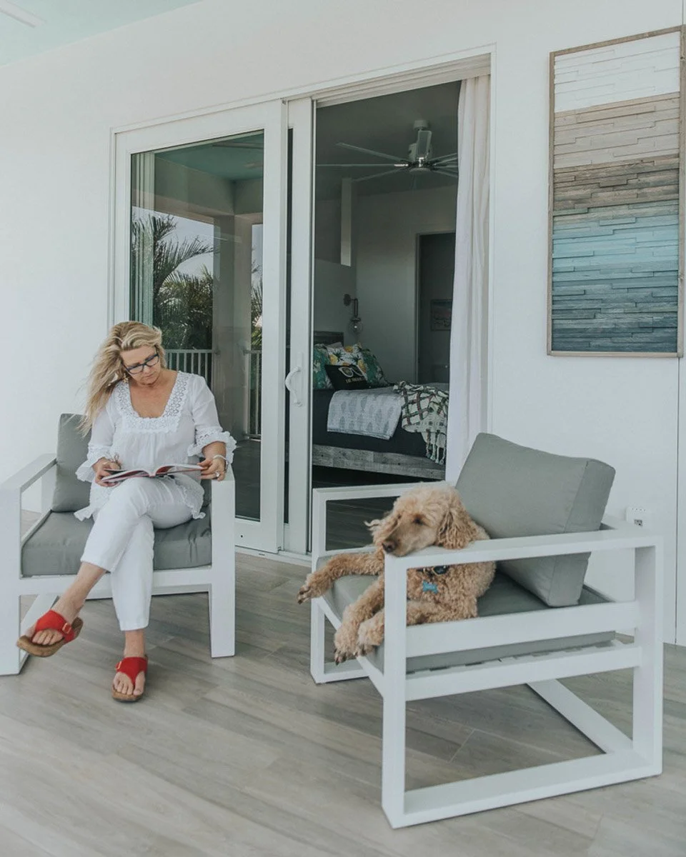 A woman with blonde hair and glasses wearing white clothing sits on a gray outdoor chair reading a book. Next to her, a golden doodle dog lies on another gray outdoor chair. Behind them is a sliding glass door leading into a bedroom.