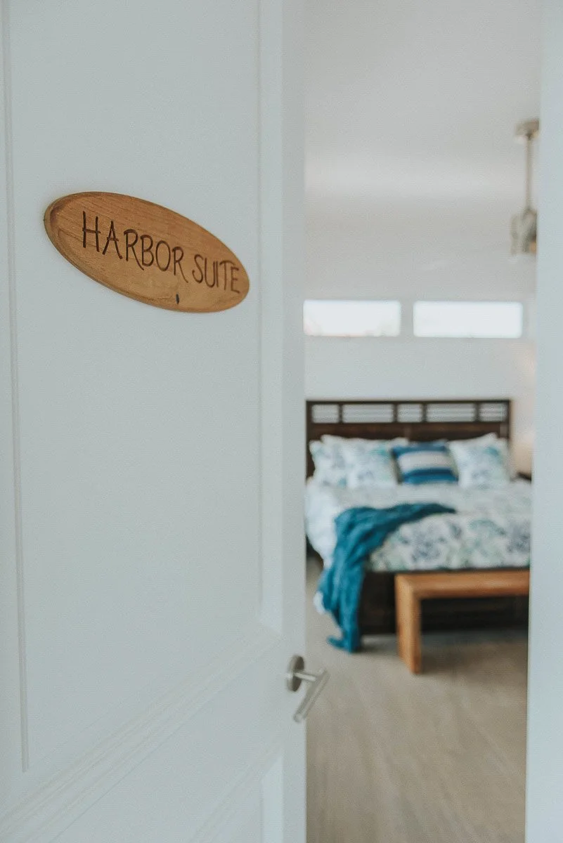 A hotel room with a wooden sign that reads 'HARBOR SUITE' on the door, which is slightly open, revealing a bed with white and blue bedding inside we can see part of the headboard and pillows.