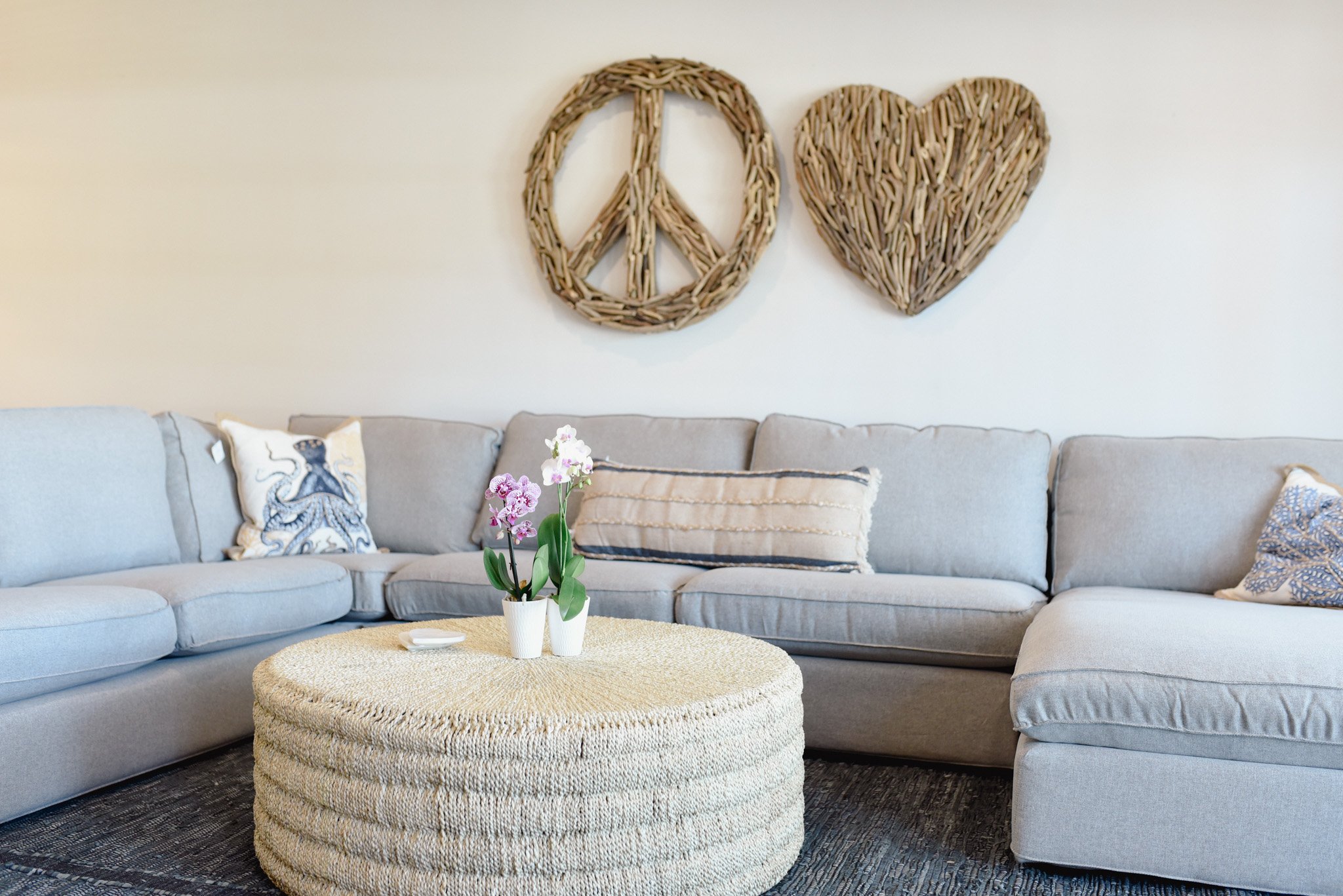Living room with a light gray sectional sofa, decorative pillows, a round woven ottoman coffee table with potted orchids, and two woven wall hangings shaped like a peace sign and a heart on the wall.
