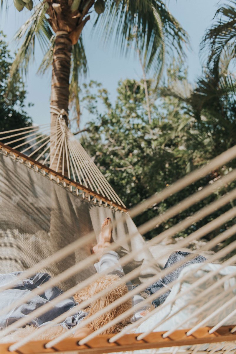 Person relaxing on a hammock outdoors, surrounded by palm trees and green foliage on a sunny day.