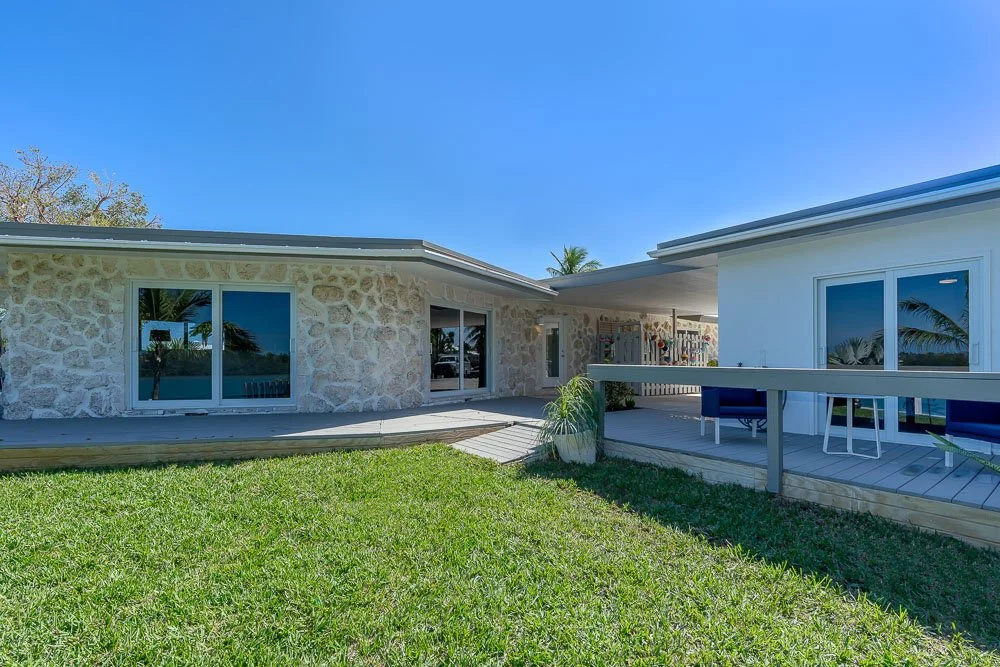 Front view of a modern house with a stone wall exterior, glass sliding doors, a wooden deck, and outdoor seating, with a grassy lawn and palm trees in the background under a clear blue sky.
