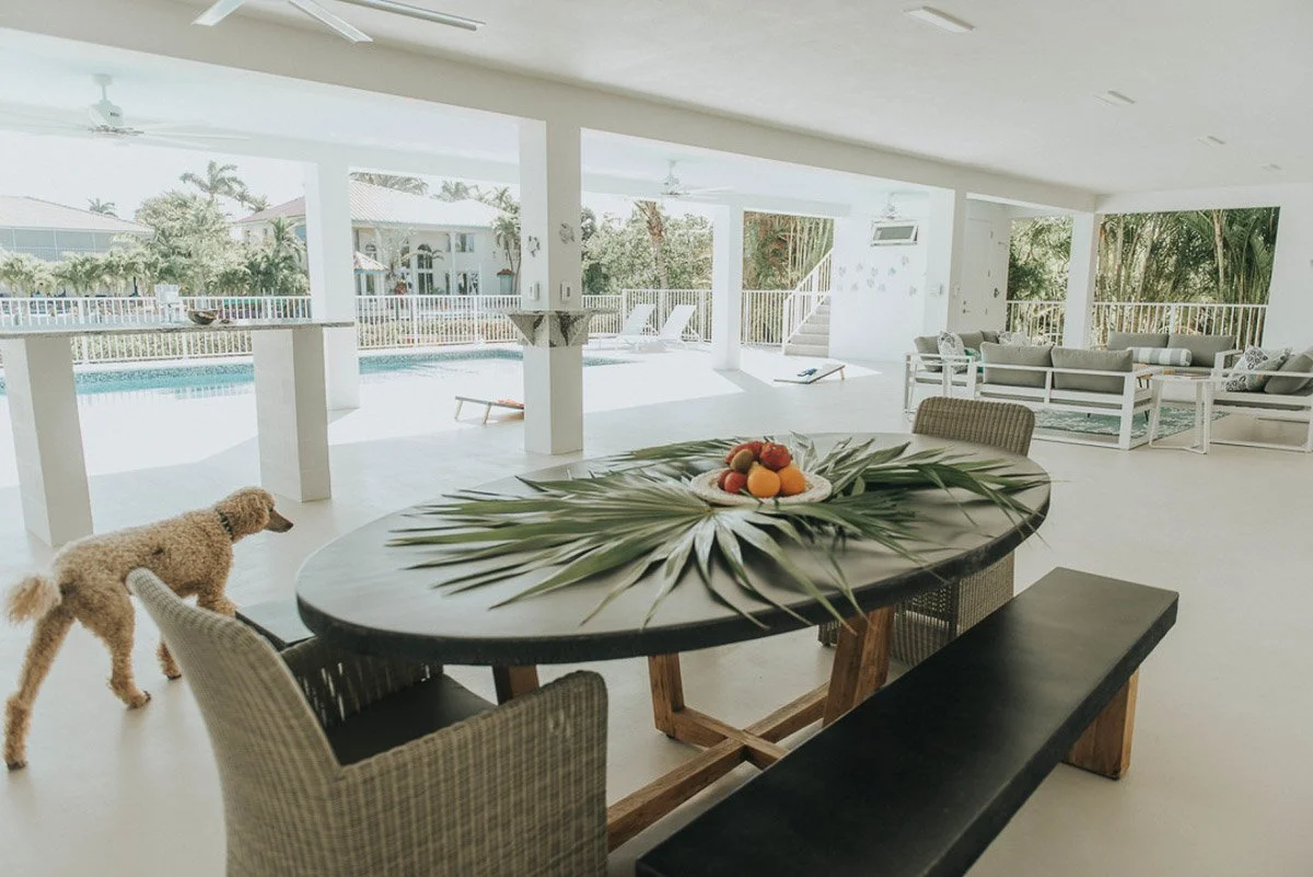 Bright, open indoor living space with a view of a swimming pool outside. A large oval table with a palm leaf centerpiece and fruit is in the foreground, surrounded by chairs and a bench. In the background, there are patio furniture and reclining chai