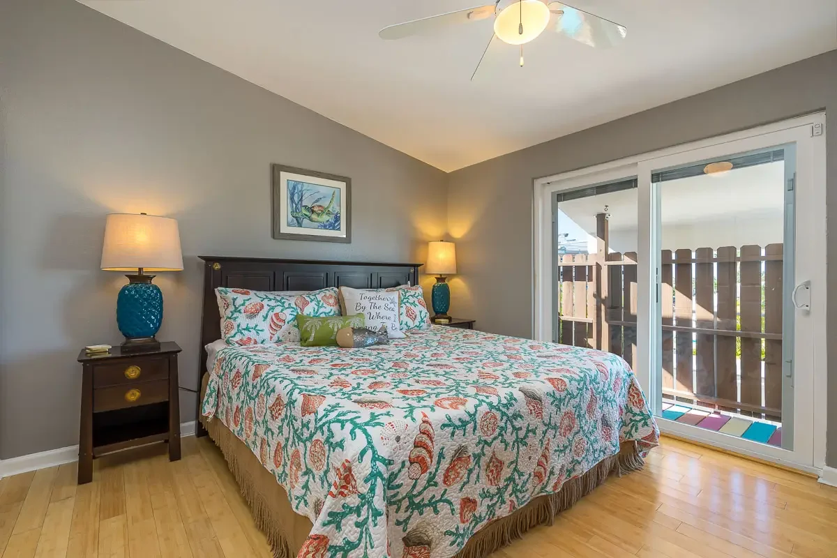 A bedroom with a bed covered in a seashell-patterned quilt, two lamps on dark wood nightstands, a framed ocean scene above the headboard, and sliding glass doors leading to a balcony.