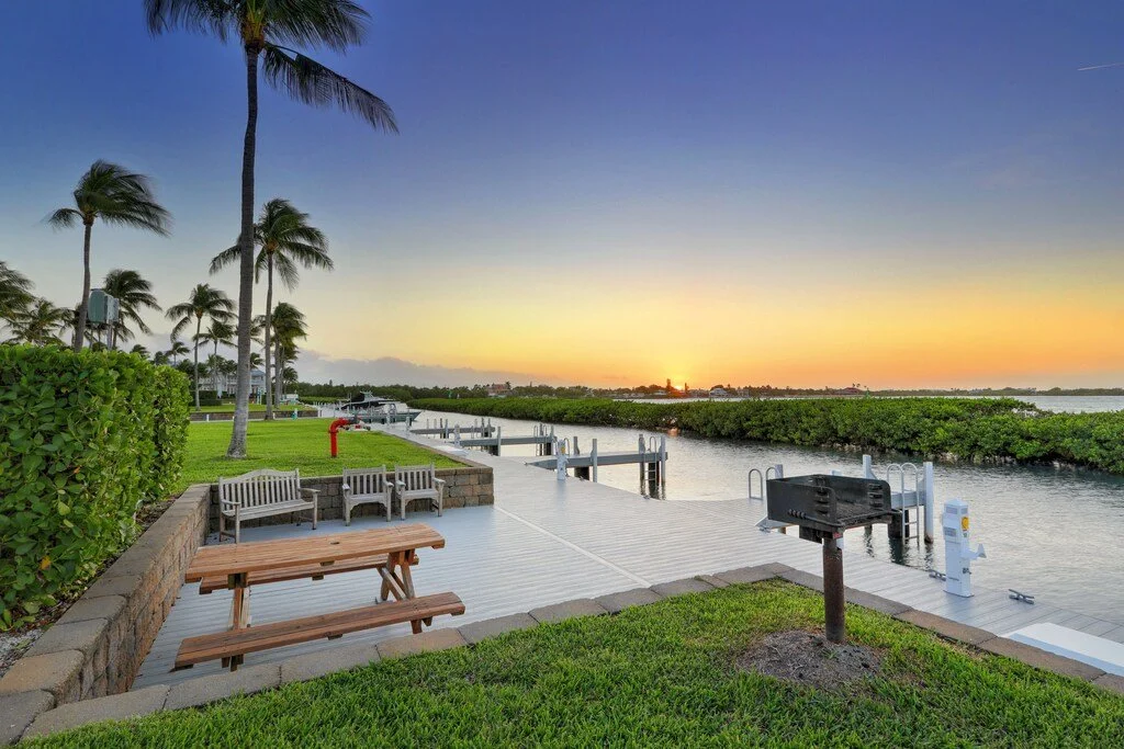 A waterfront scene at sunset with palm trees, a wooden pier, benches, a black grill, and lush green shrubbery