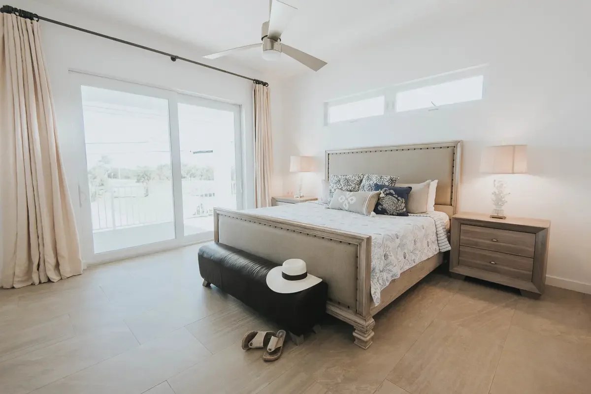 A bright bedroom with a large bed, nightstands, lamps, and a sliding glass door leading outside. A hat and sandals are on a bench at the foot of the bed.