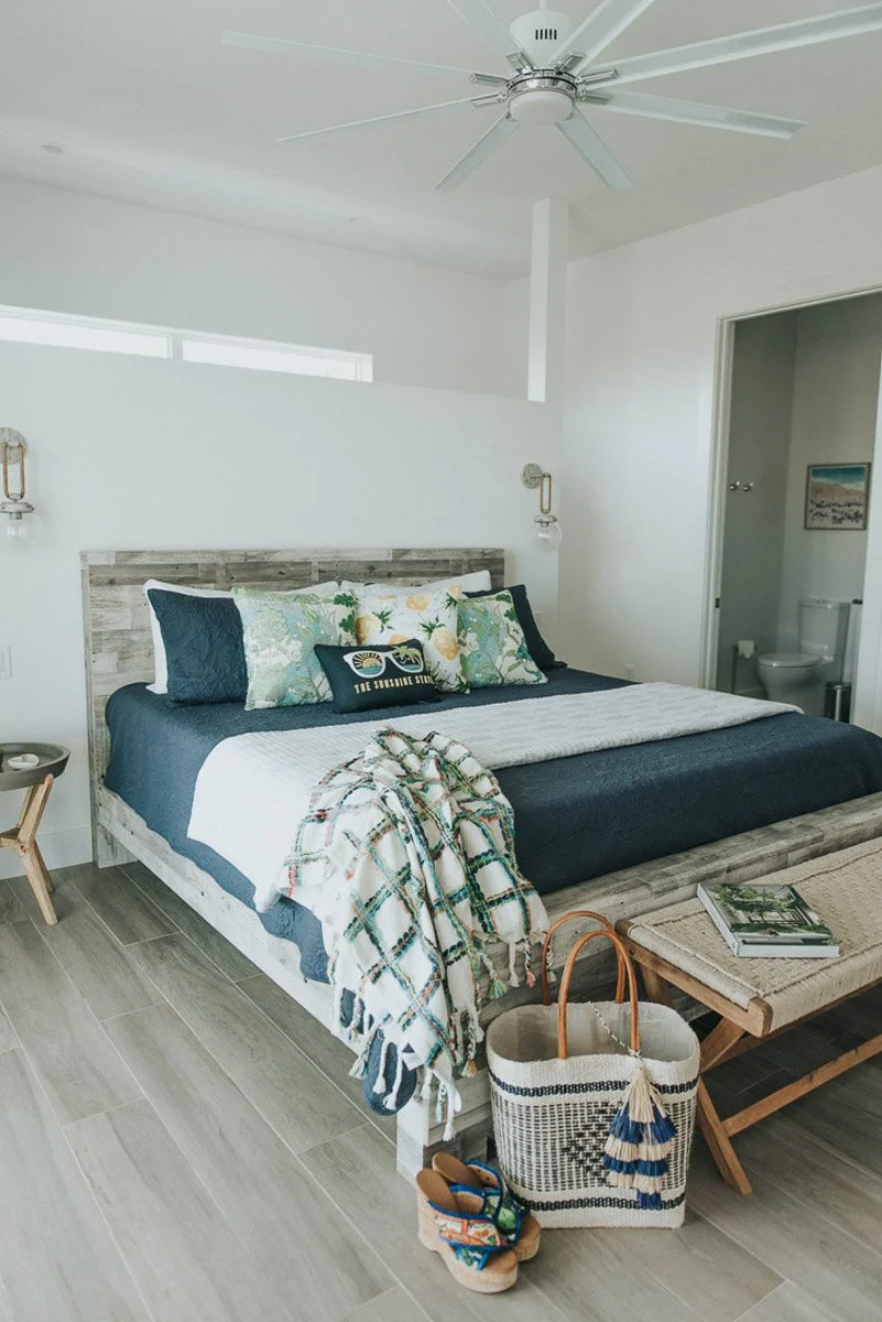 A bedroom with a reclaimed wood bed frame, navy bedding, pillows with pineapple and floral prints, and a throw blanket. There are wall-mounted lights, a small side table, a woven bench with books, a tote bag, and sandals. The room has a ceiling fan a