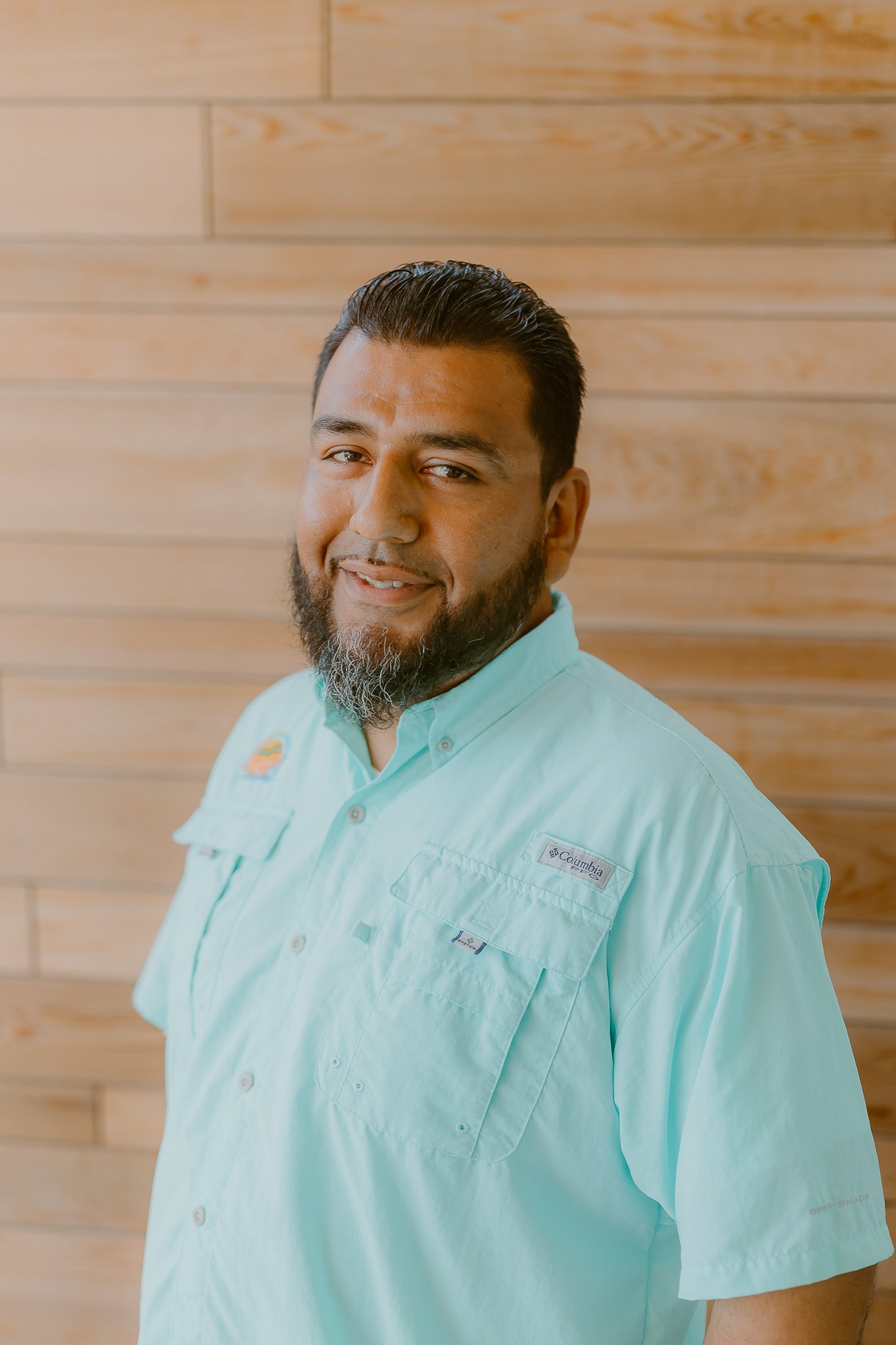 Man with dark hair and beard, wearing a light blue Columbia shirt, standing against a wooden wall.
