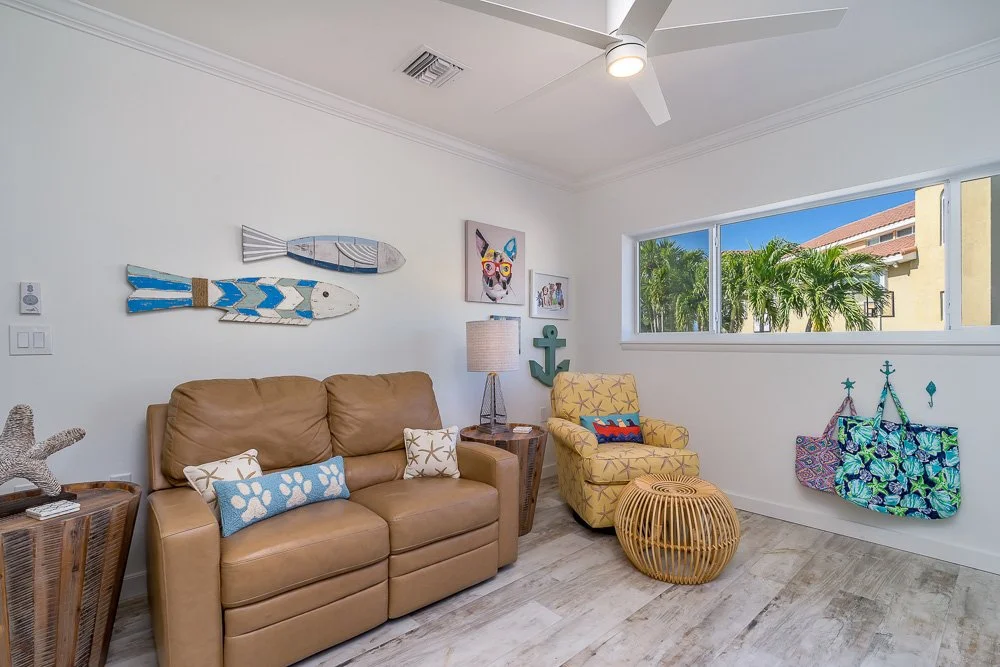 Living room with a tan leather sofa, yellow armchair, and beach-themed decor including wooden fish wall art, starfish pillow, and a hanging tote bag with tropical print. Large window showing palm trees and a building outside.