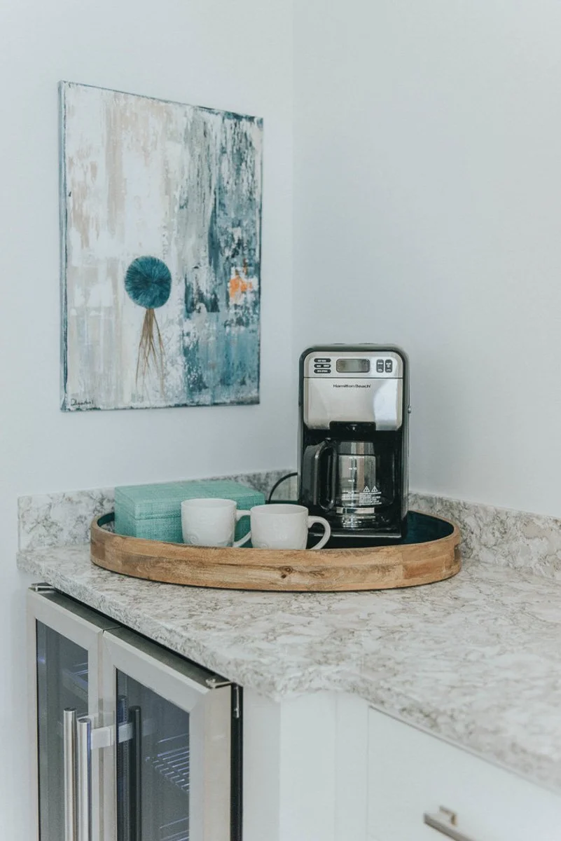 Coffee maker with two white mugs on a round wooden tray on a marble countertop, decorated with a teal napkin, next to a small wine fridge, with a colorful abstract painting on the wall behind.