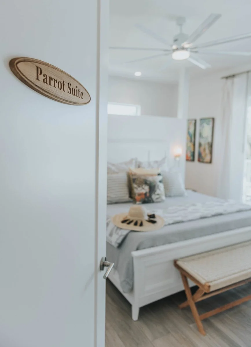 Open door with a sign that says 'Parrot Suite,' revealing a bright bedroom with white furniture, artwork, and a ceiling fan.