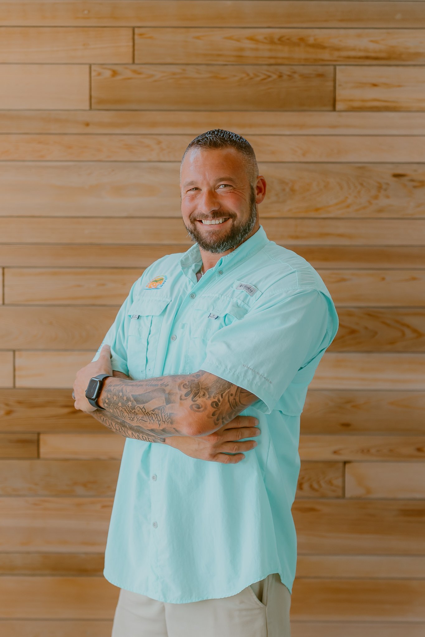 A man with a beard, tattoos, and a smartwatch smiling with arms crossed against a wooden wall background.