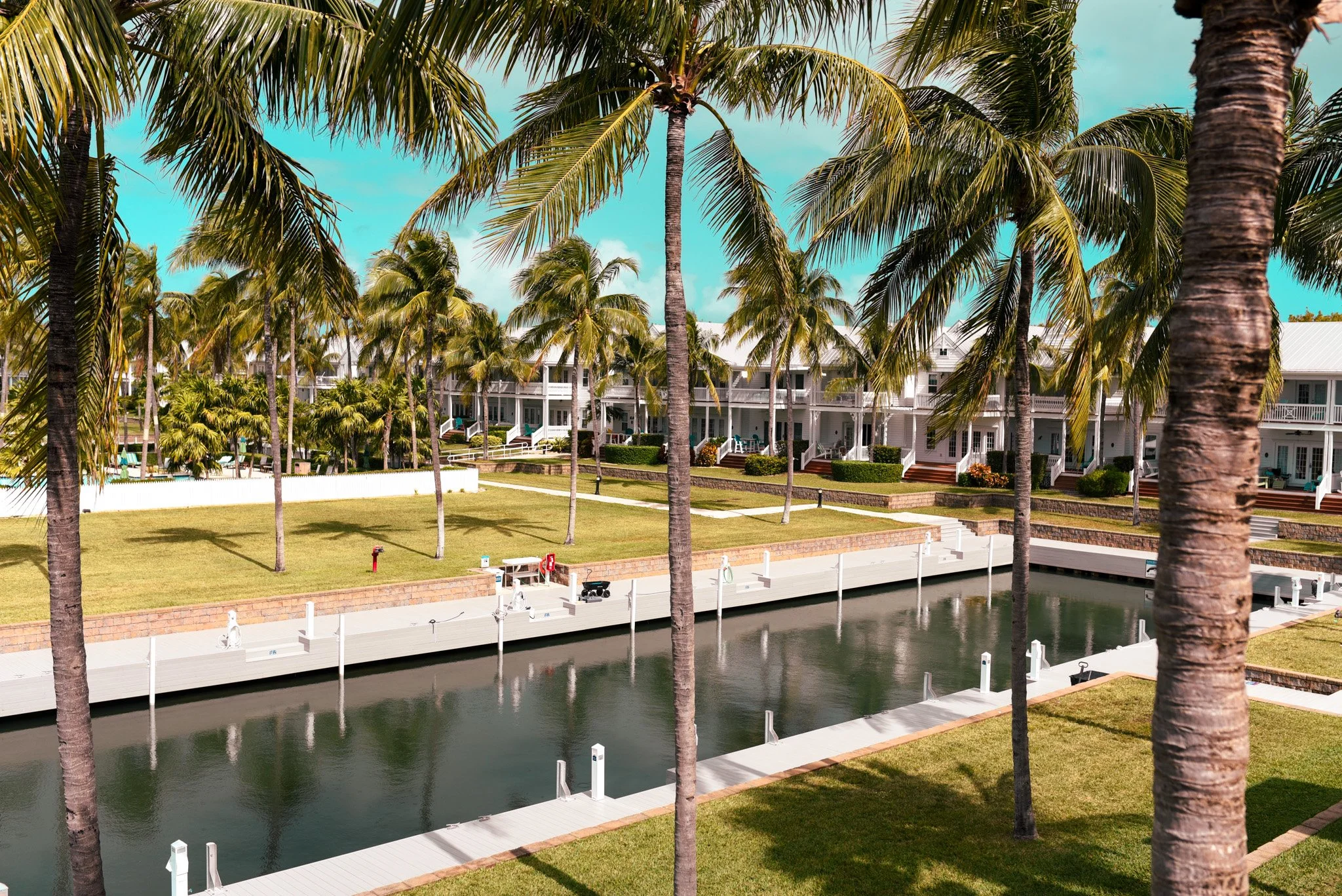 A canal with a dock and palm trees in front of a multi-story white resort or hotel with balconies, under a partly cloudy sky.