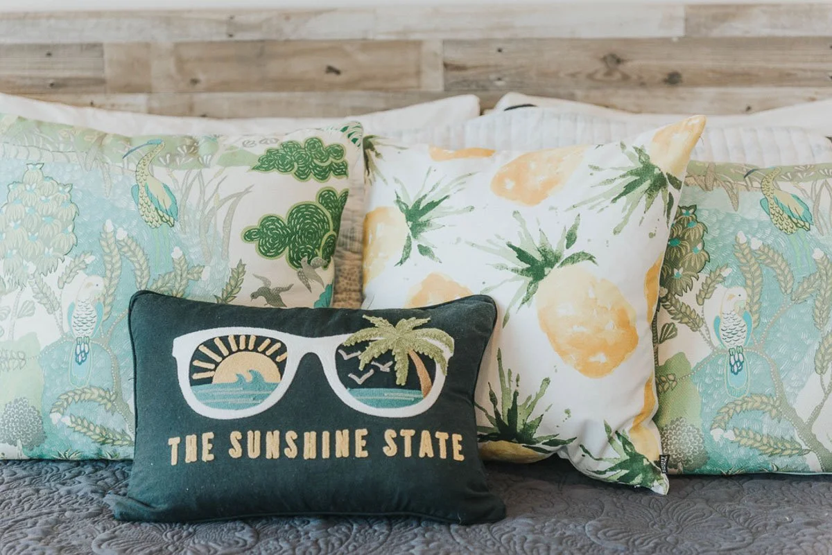 Decorative pillows on a bed with a wooden headboard. The pillows have tropical and beach-themed patterns, including pineapples, parrots, and palm trees. The front pillow has a design with sunglasses, a palm tree, and the text "The Sunshine State."