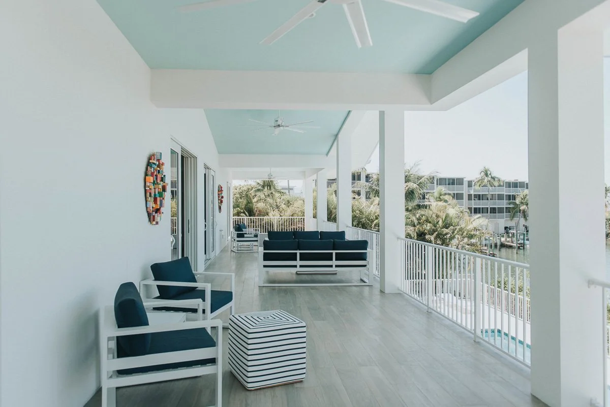 Bright, spacious balcony with white walls and railings, featuring navy and white furniture, artwork on the wall, ceiling fans, and views of palm trees and neighboring buildings.