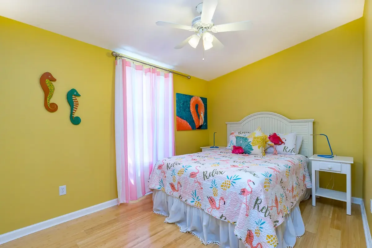 A bedroom with yellow walls, a white bed with flamingo and pineapple themed bedding, pink and white curtains, wall decorations including seahorse art and a flamingo painting, a ceiling fan with lights, and two white bedside tables with blue lamps.