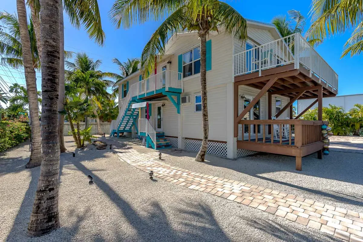 Two-story beach house with a white exterior, surrounded by palm trees, with blue and brown wooden stairs and balconies, set on a sandy landscape with a brick pathway.