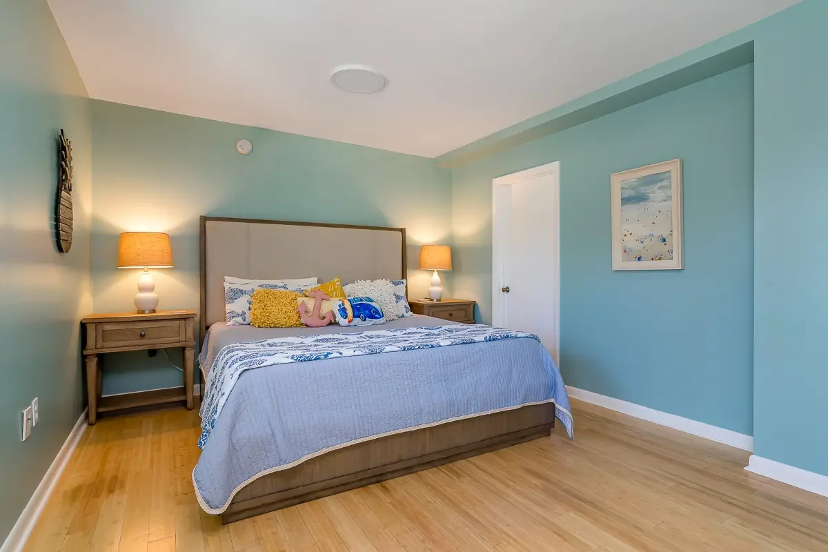 A bedroom with a large bed, light blue walls, and wooden nightstands with lamps. The bed has decorative pillows and toys, and there is a framed picture on the wall.