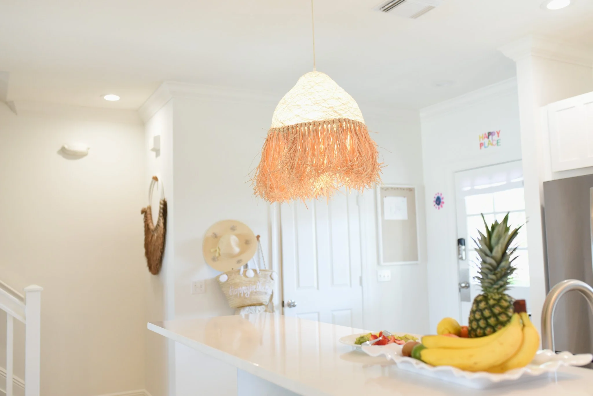 Kitchen with a hanging lampshade, fruit on a white plate, and a door with a window. Decor includes a hat and a woven wall hanging.