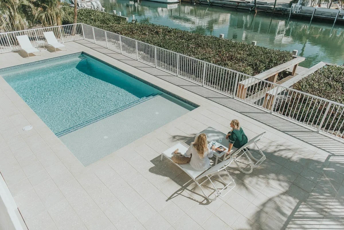 Two women relaxing on poolside lounge chairs near a swimming pool with a marina in the background.
