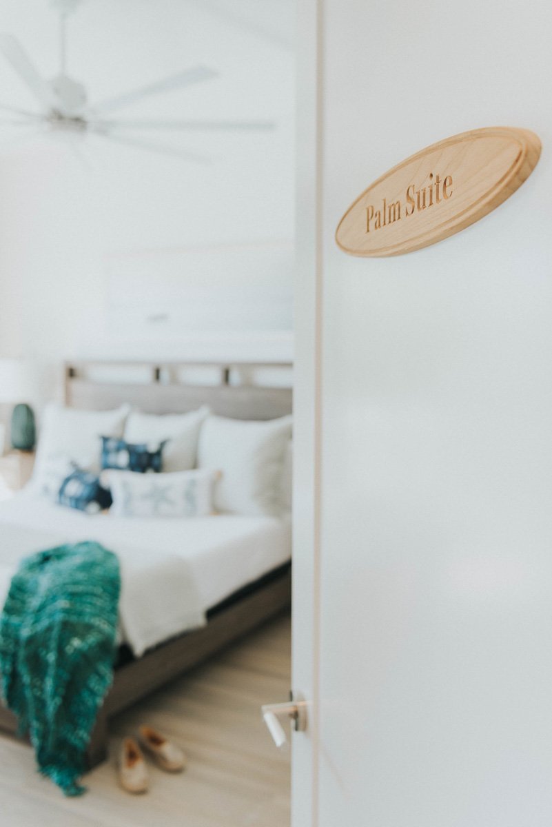 A hotel room with a bed, decorated pillows, a green blanket, slippers on the floor, and a wooden sign on the door that says "Palm Suite."