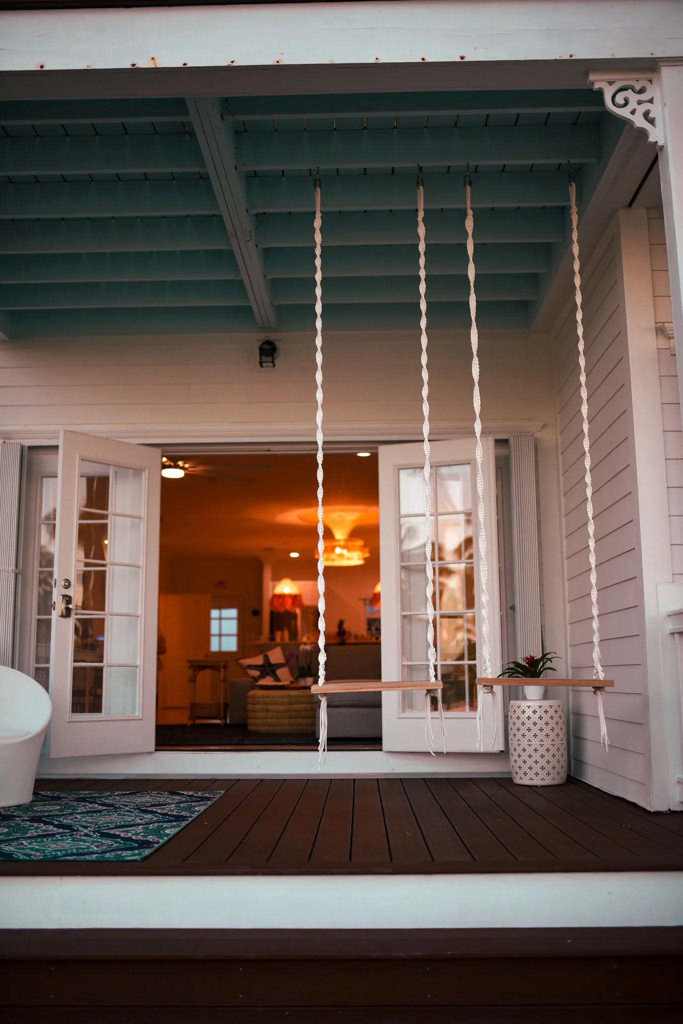 View of a porch with a ceiling painted in teal, featuring four rope swings hanging from hooks. The porch has white siding, and through open French doors, a living room with warm lighting, a sofa with pillows, and a chandelier are visible inside.