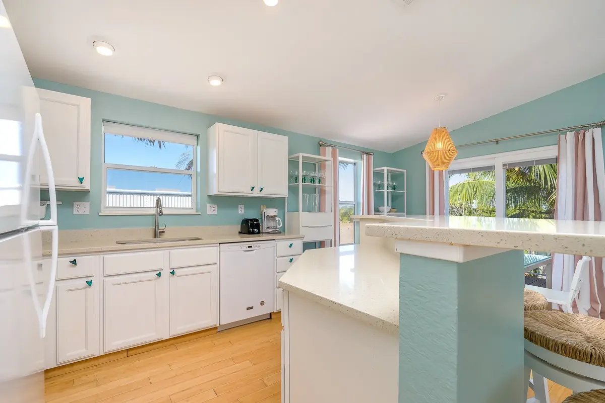 Bright kitchen with white cabinets, light wood flooring, and large windows showing palm trees outside. There are small teal accents on cabinet knobs and a white countertop with a breakfast bar. A hanging pendant light is also visible.