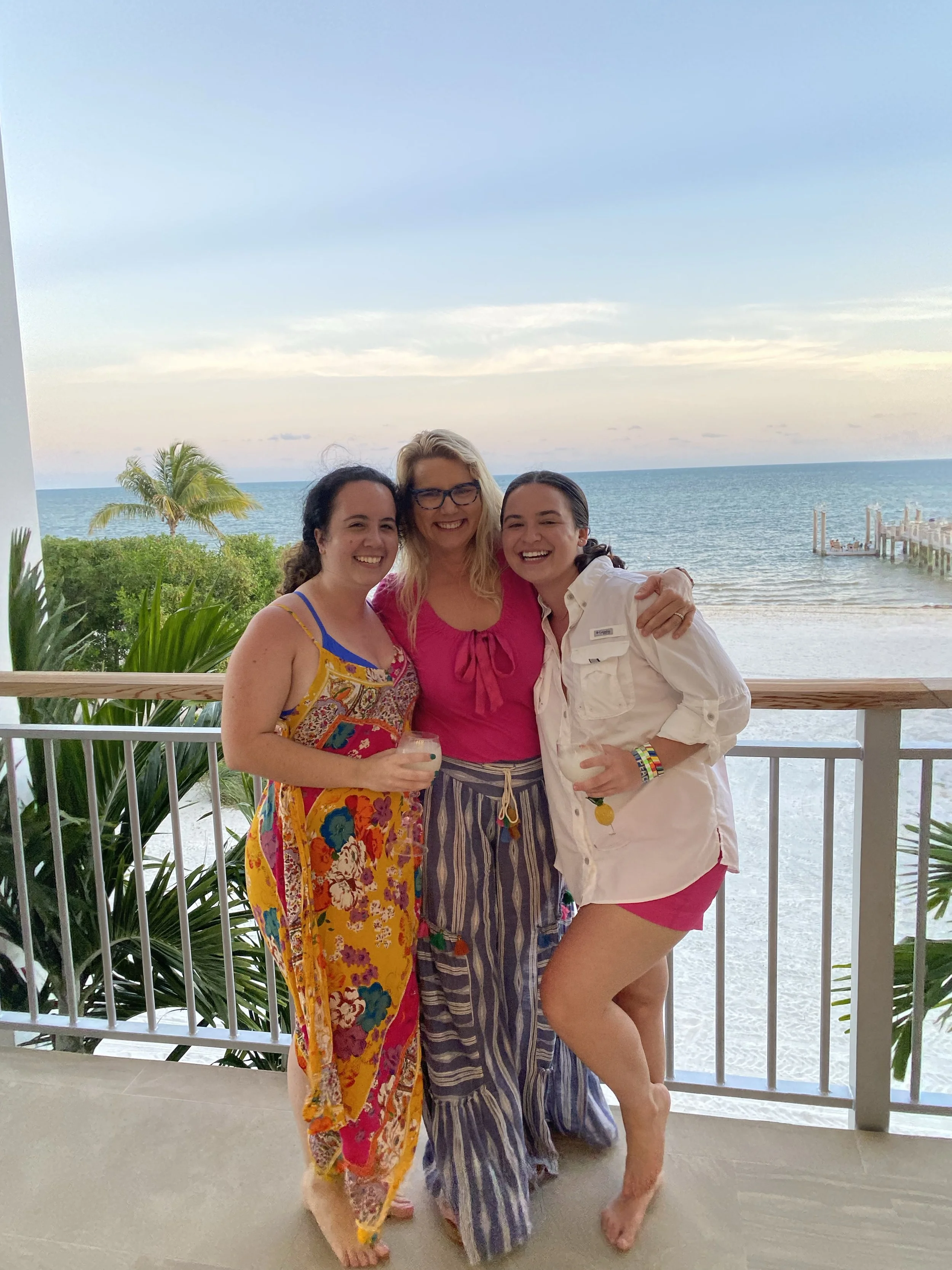 Three women smiling, embracing, and holding drinks on a balcony overlooking a beach and ocean, with a cloudy sky and palm trees in the background.