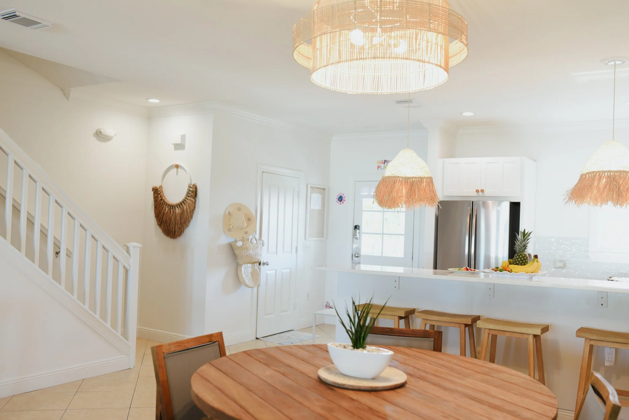 Bright, modern kitchen with white cabinets and stainless steel refrigerator, complemented by a round wooden dining table with chairs, decorative hanging lights, and fruit on the counter.
