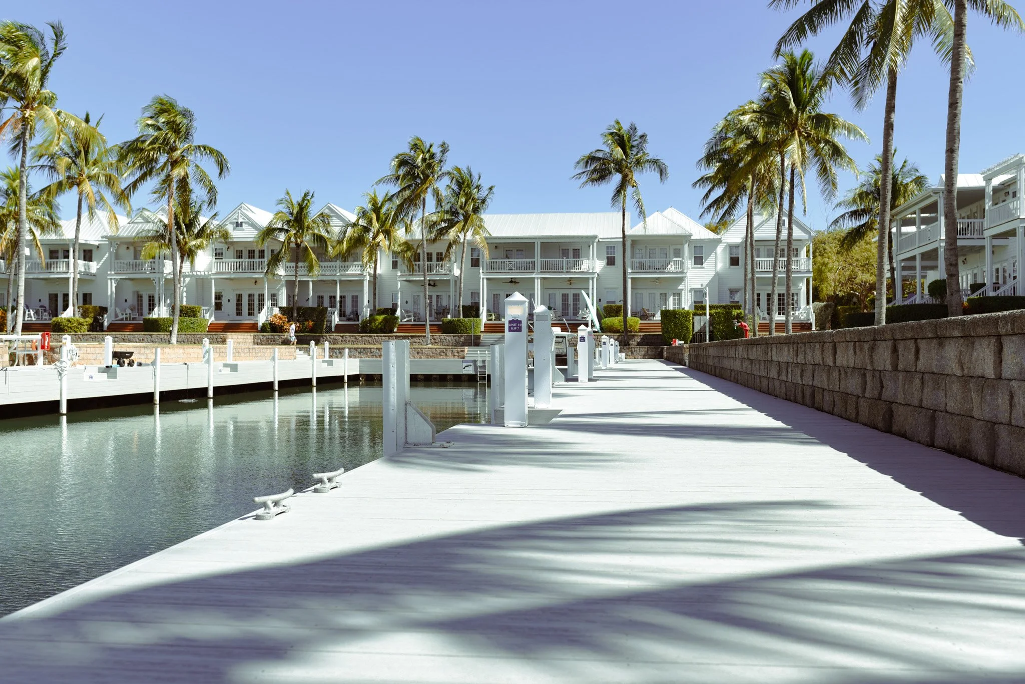 A waterfront view of white residential buildings with porches and balconies, surrounded by palm trees under a clear blue sky. A white dock extends over calm water in the foreground.