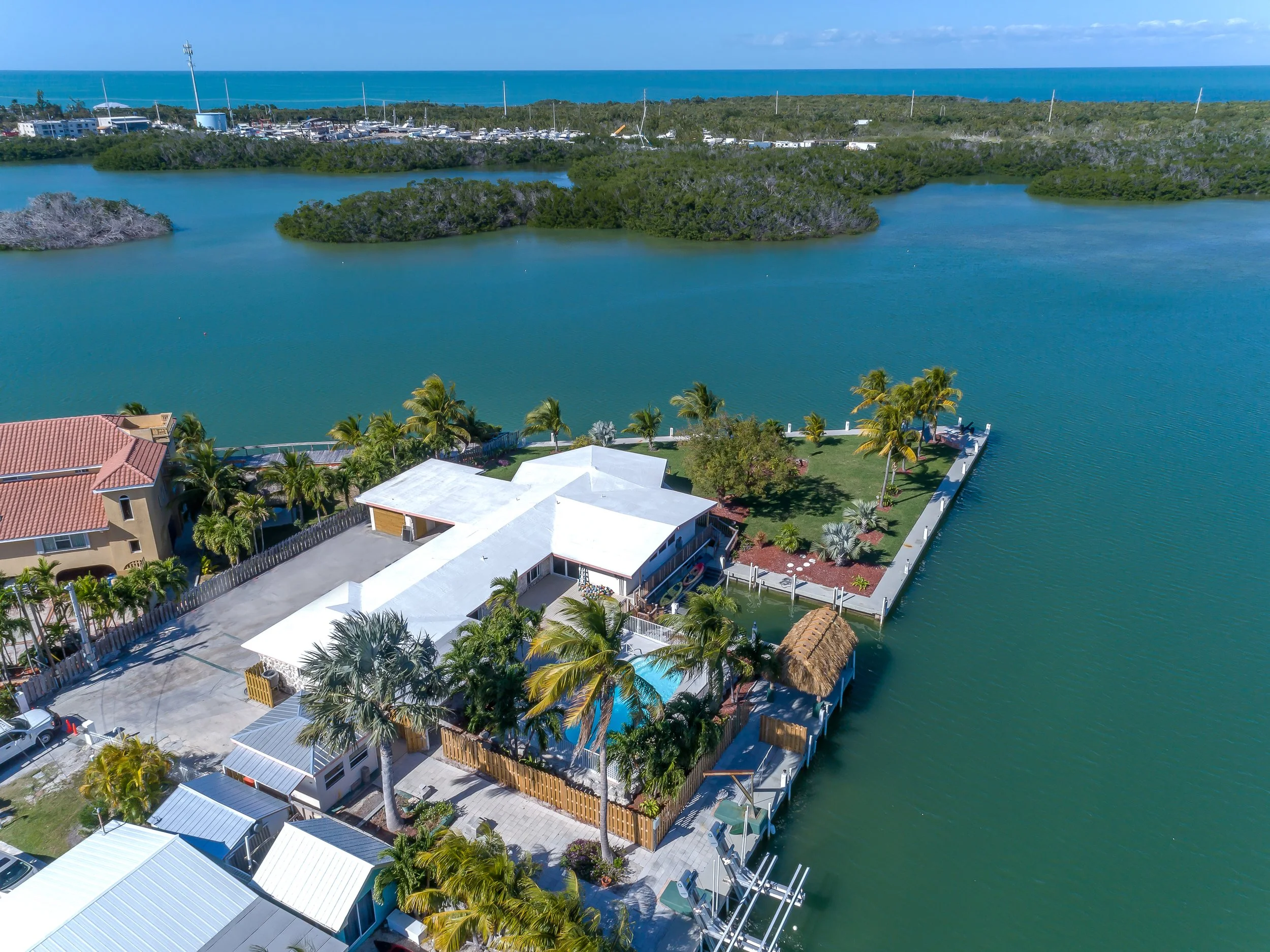 Aerial view of a waterfront house with a boat dock, swimming pool, palm trees, and a lawn surrounded by water, with a bridge and open water in the background.