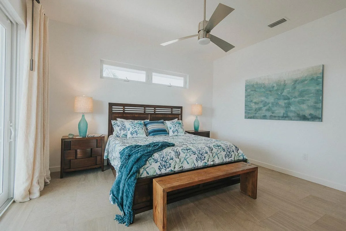 A bedroom with a wooden bedframe, white bedding with blue and white floral patterns, and a blue throw blanket. There are two matching teal lamps on bedside tables, a wooden bench at the foot of the bed, a large abstract painting on the white wall, an