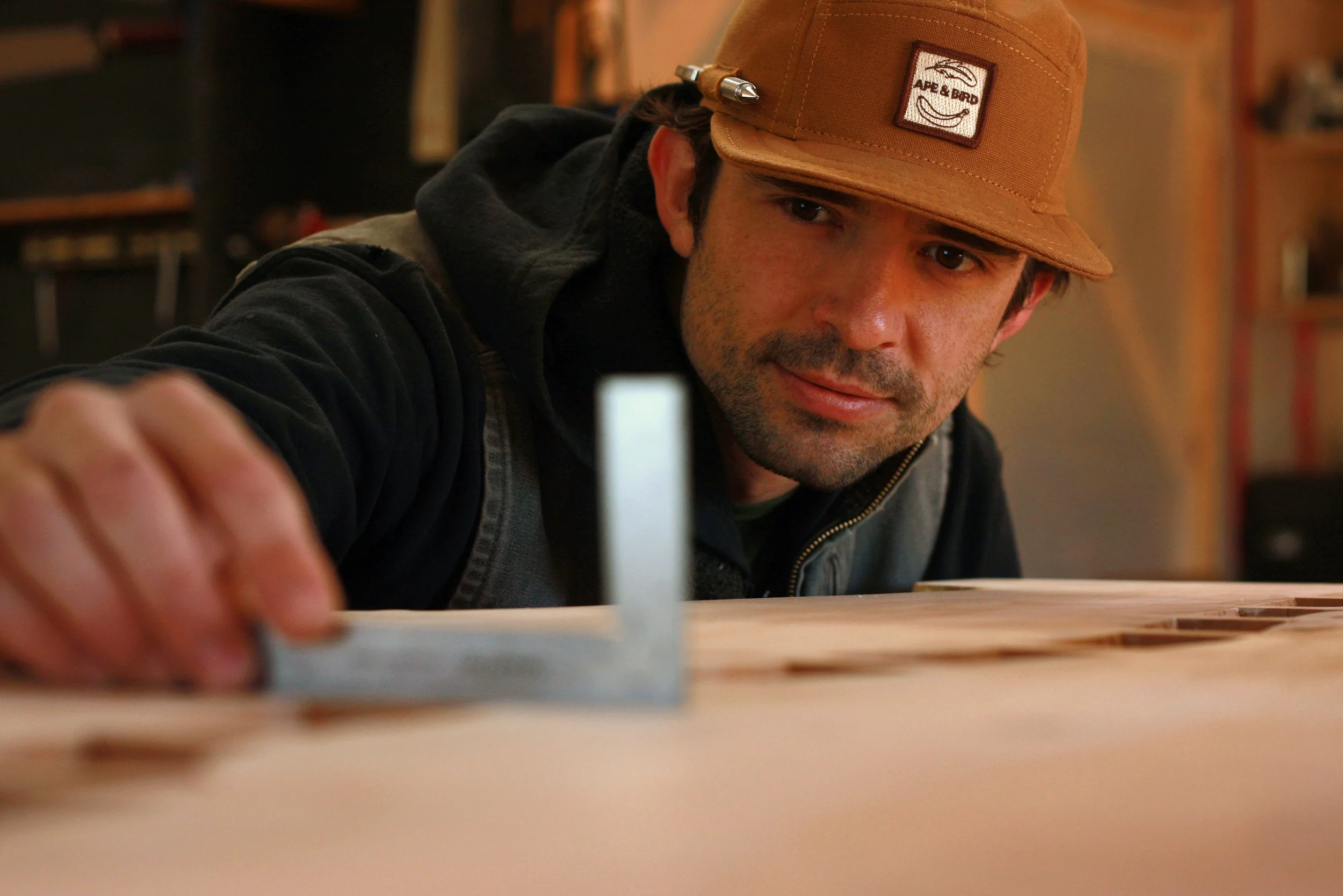 A man wearing a tan cap with a patch that says 'APE & BIRD' and a black hoodie, as he leans over a workbench, holding a small rectangular object in focus.