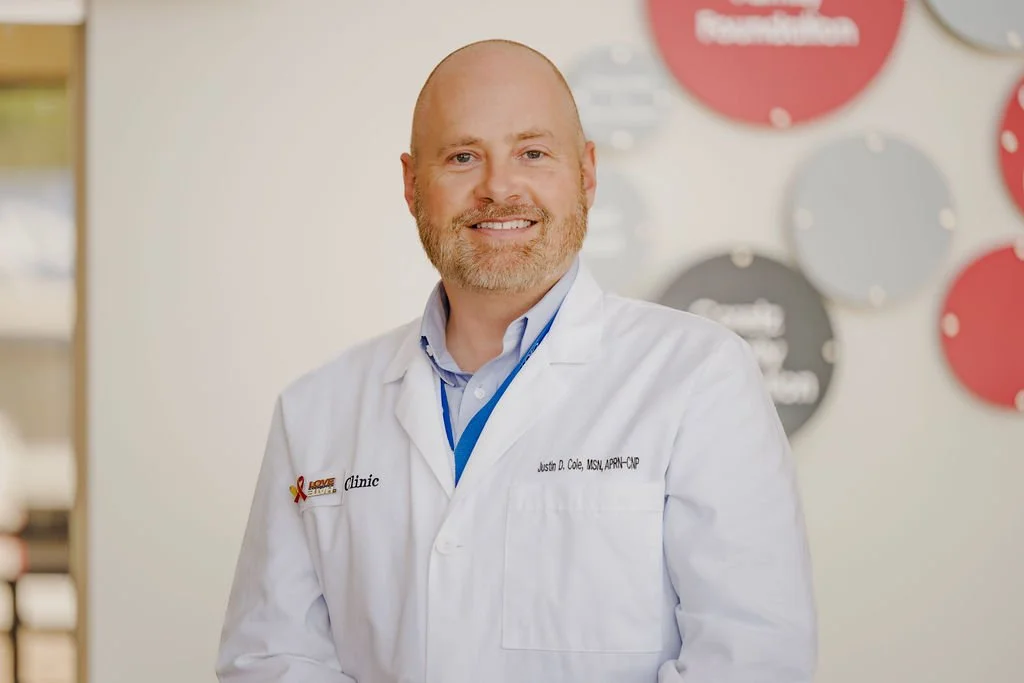 A male doctor with a beard and short hair smiling, wearing a white lab coat with a name tag, standing in front of a wall with red and gray circular signs.