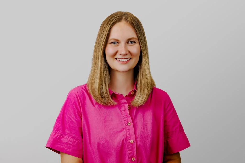 A young woman with shoulder-length blonde hair, smiling, wearing a bright pink button-up shirt against a plain light gray background.