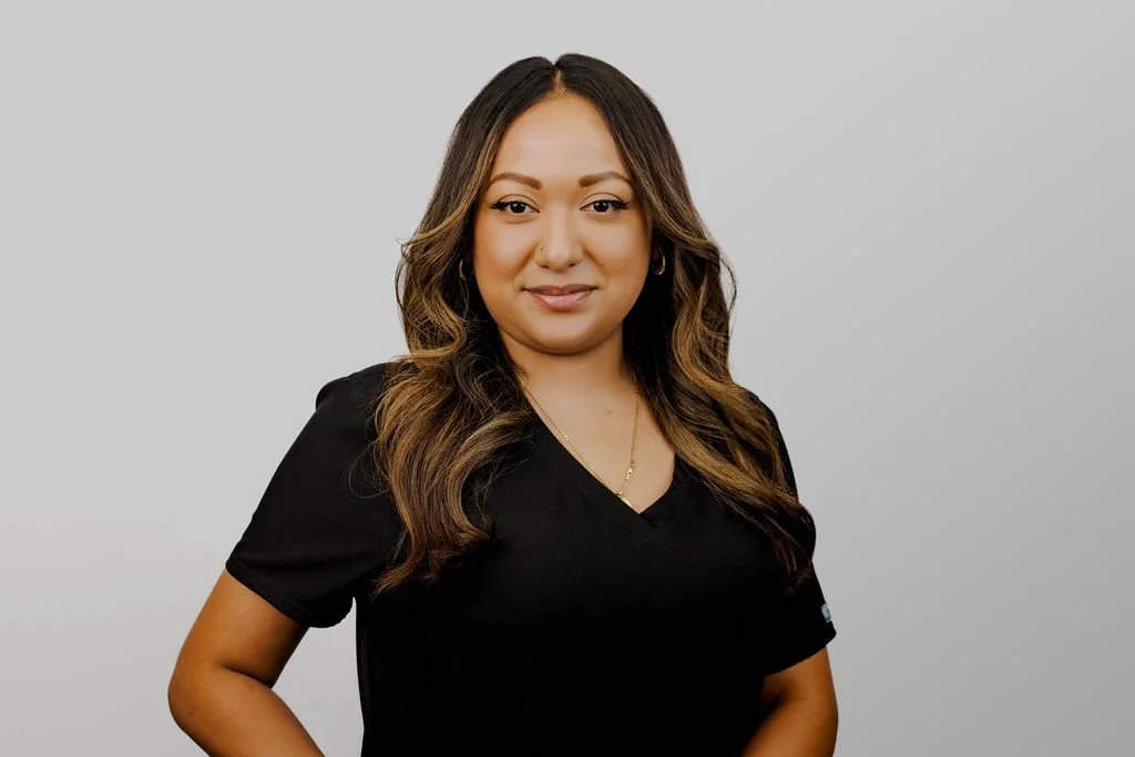 A woman with wavy brown hair wearing a black top, standing against a light gray background.