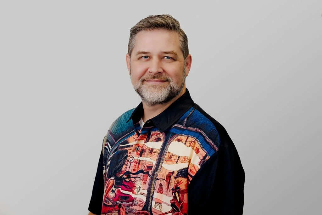 A smiling man with short gray hair and a beard, wearing a colorful, patterned, short-sleeved shirt over a black jacket, standing against a plain light gray background.