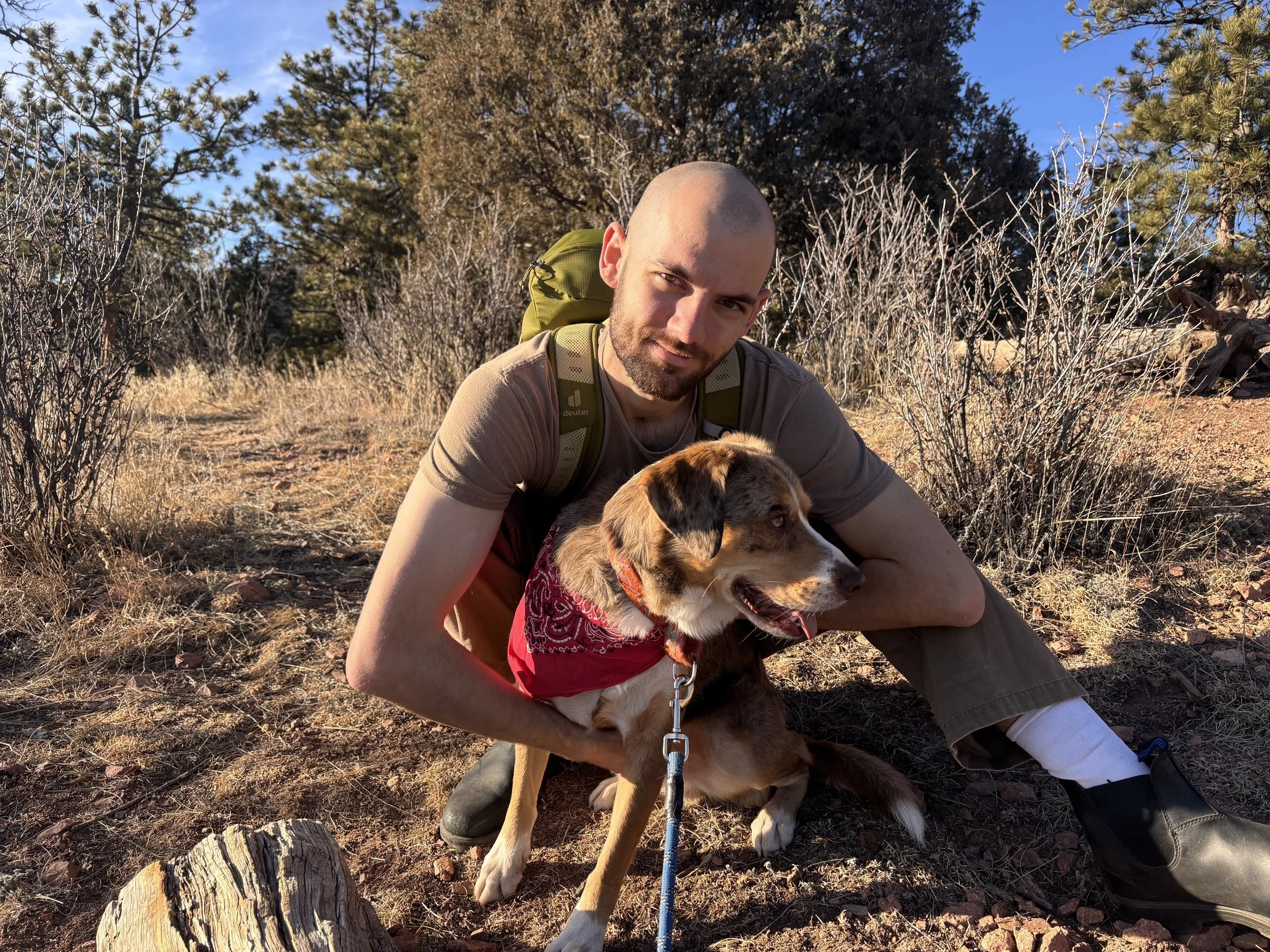 A man with a shaved head and beard crouching on a dirt trail in a forest, holding a brown and white dog with a red bandana, against a background of trees and blue sky.