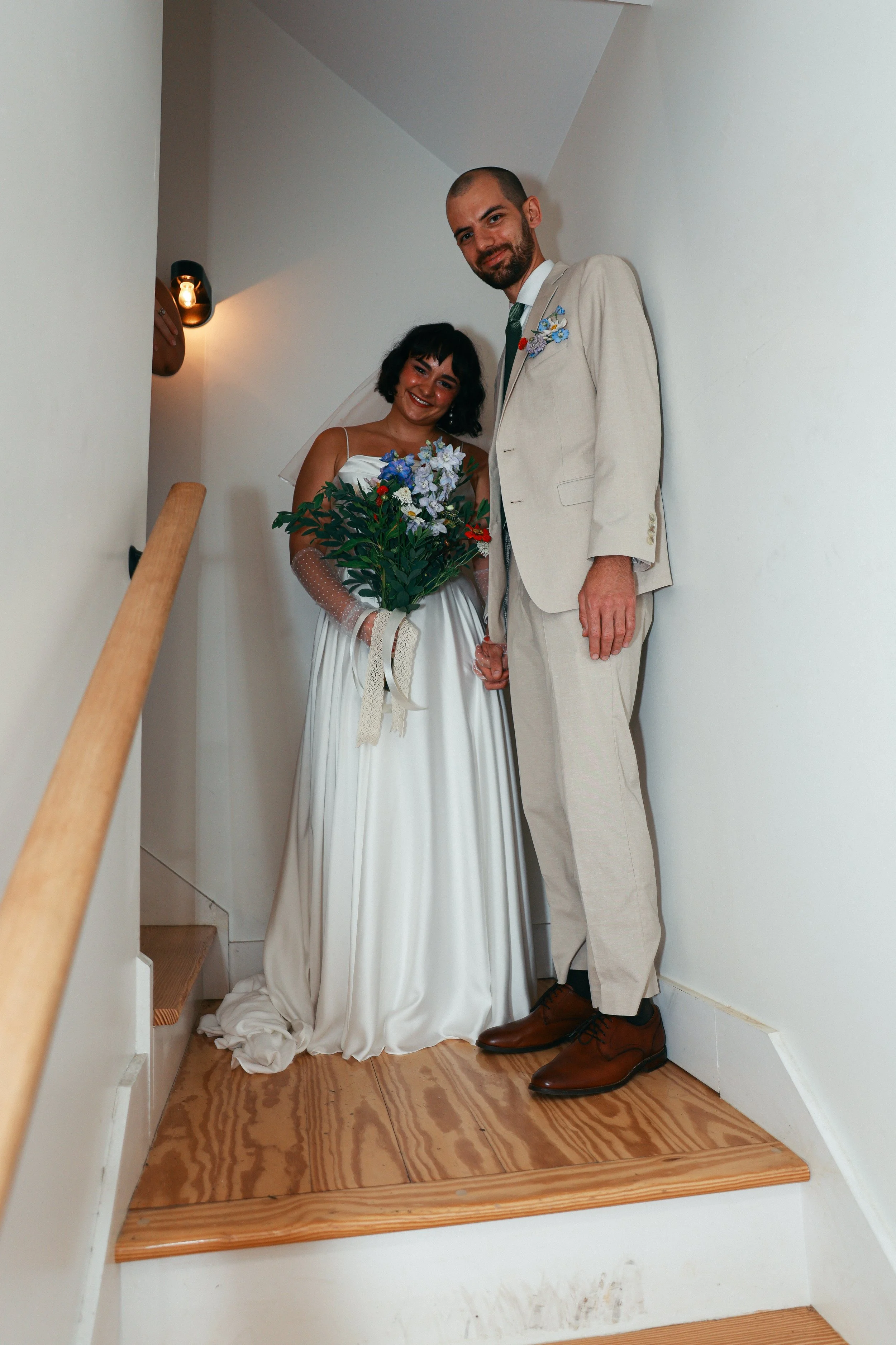 A bride in a white wedding dress holding a bouquet of flowers, and a groom in a light-colored suit holding hands, standing in a small enclosed space.